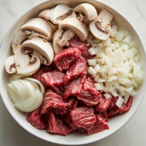 A bowl containing raw ingredients for beef stroganoff - chunks of beef, sliced mushrooms, and chopped onions ready for cooking.