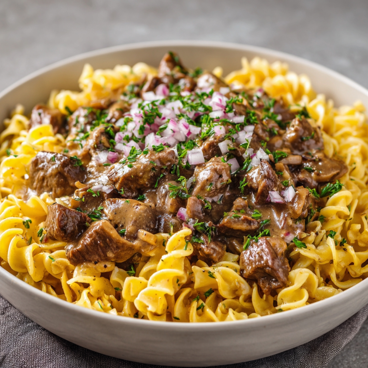 A bowl of beef stroganoff served over curly egg noodles, topped with creamy mushroom sauce, chopped onions, and fresh parsley.