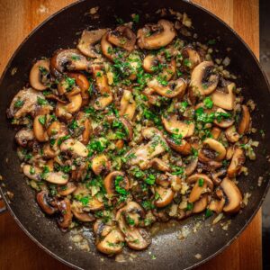 A sauté pan with sliced mushrooms, onions, and herbs being cooked, ready for the Beef Wellington.
