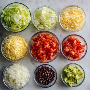 An assortment of burrito bowl ingredients in small glass bowls, including shredded lettuce, shredded cheese, diced tomatoes, chopped onions, black beans, and mashed avocado on a marble surface.
