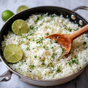 A skillet of cilantro lime rice garnished with lime halves and chopped cilantro, with a wooden spoon resting in it.