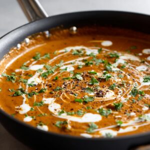 A pan filled with rich, creamy Indian curry sauce, garnished with fresh cilantro and drizzled with cream, showing a swirl pattern on top.
