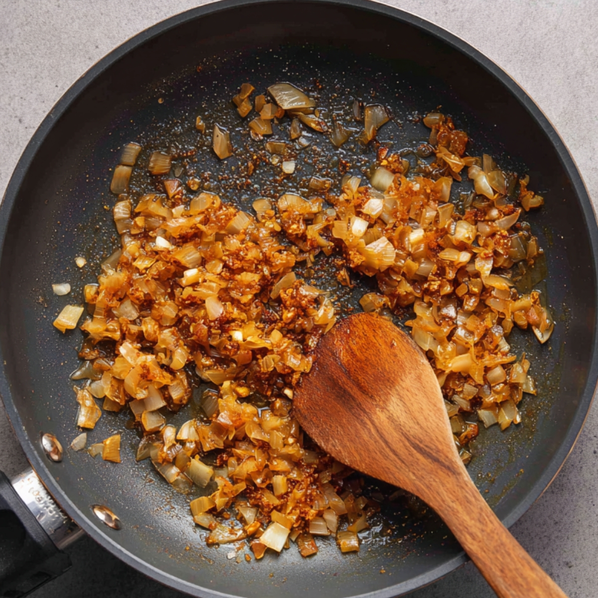 A frying pan with sautéed onions, garlic, and spices being cooked until golden brown, stirred with a wooden spoon.