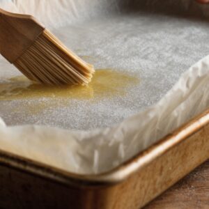 A baking tray lined with parchment paper being brushed with melted butter, preparing it for baking.