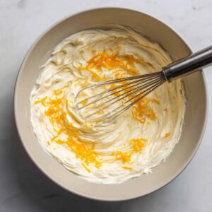 A mixing bowl filled with creamy cheesecake batter topped with fresh orange zest, and a whisk resting inside.