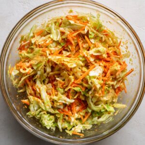 A clear glass bowl filled with a colorful coleslaw made of shredded cabbage and carrots, coated in a spicy reddish dressing.