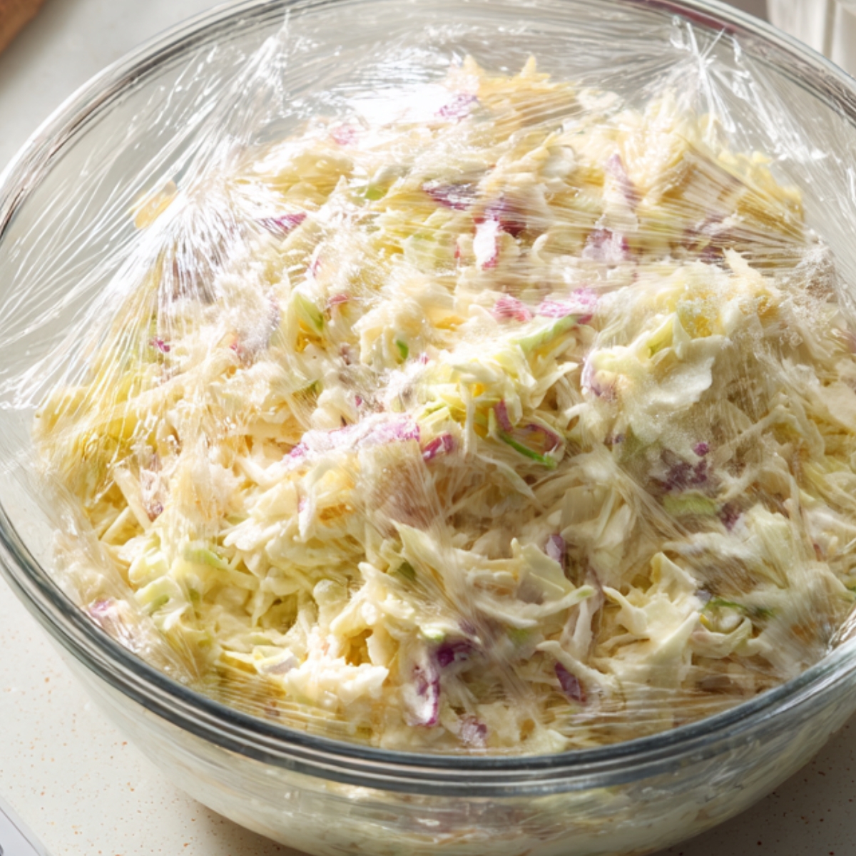 A glass bowl of creamy coleslaw covered tightly with plastic wrap, showing shredded cabbage and carrots mixed with dressing.