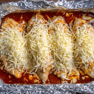Unbaked chicken breasts in a foil-lined tray, covered in red sauce and topped generously with shredded cheese.