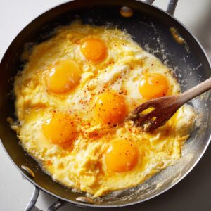 A frying pan with several eggs being scrambled, with bright yellow yolks visible and a sprinkle of seasoning in the process.