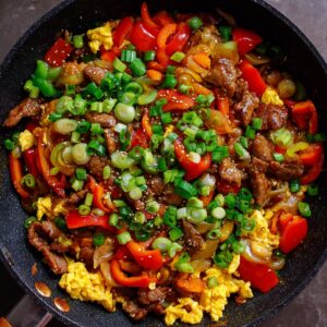 A vibrant stir-fry with scrambled eggs, slices of red and yellow bell peppers, and green onions, garnished with sesame seeds in a black frying pan.