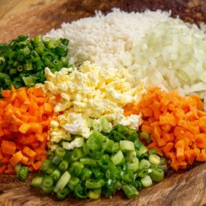 A collection of diced vegetables and cooked rice neatly arranged on a wooden surface. The ingredients include onions, green onions, carrots, scrambled eggs, and rice, ready for cooking.