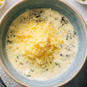 A bowl of mixed ingredients for a frittata, including shredded cheese, milk, and fresh herbs, ready to be whisked together.