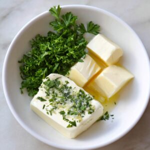 A white bowl containing softened butter, chopped parsley, and minced garlic being prepared for garlic bread spread.