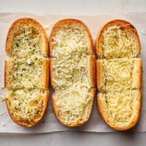 Three sliced bread halves on parchment paper, topped with garlic butter and shredded cheese, ready to be baked.