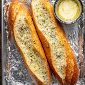 Two baguettes on a foil-lined tray, slathered with garlic butter and sprinkled with herbs and cheese before baking.
