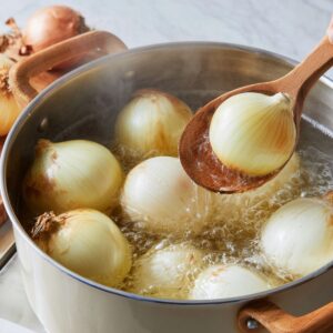 Whole onions boiling in a pot of water, with one being lifted out using a wooden spoon during preparation for stuffing.