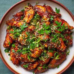 A plate of golden-brown honey garlic chicken garnished with chopped parsley, sliced green onions, and sprinkled with sesame seeds. The chicken pieces are glazed in a shiny, sticky sauce.