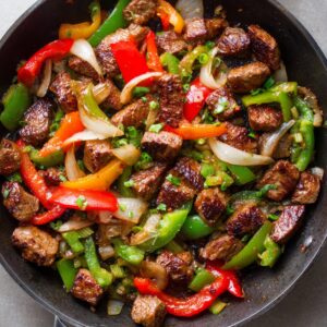 A skillet with colorful bell peppers (red, green, and yellow), onions, and browned chunks of beef. The dish appears freshly cooked with vibrant colors and a slight char on the beef.