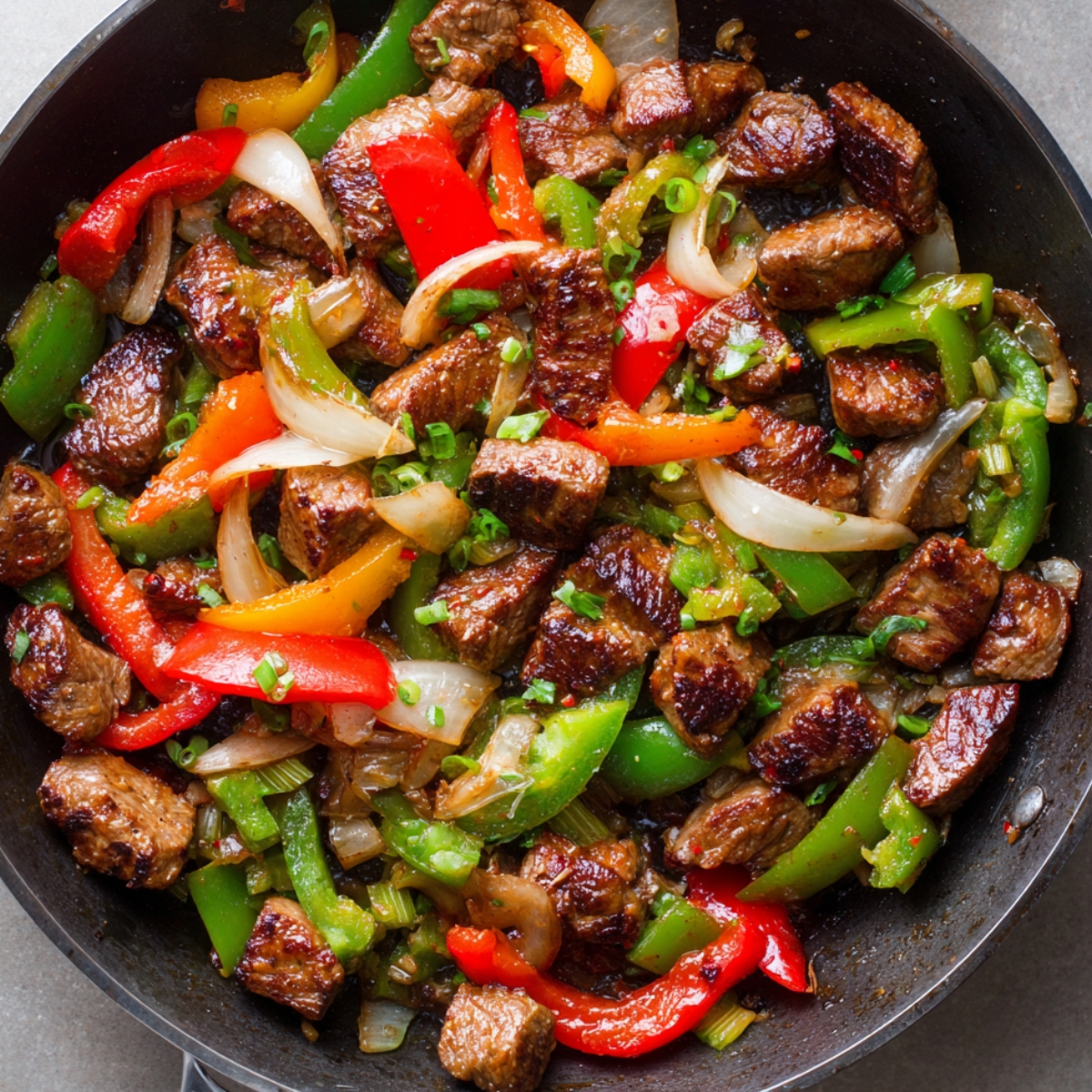 A skillet with colorful bell peppers (red, green, and yellow), onions, and browned chunks of beef. The dish appears freshly cooked with vibrant colors and a slight char on the beef.