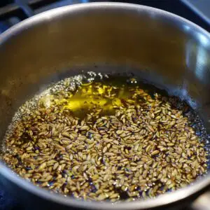 A close-up of fennel seeds frying in hot oil inside a metal pot.