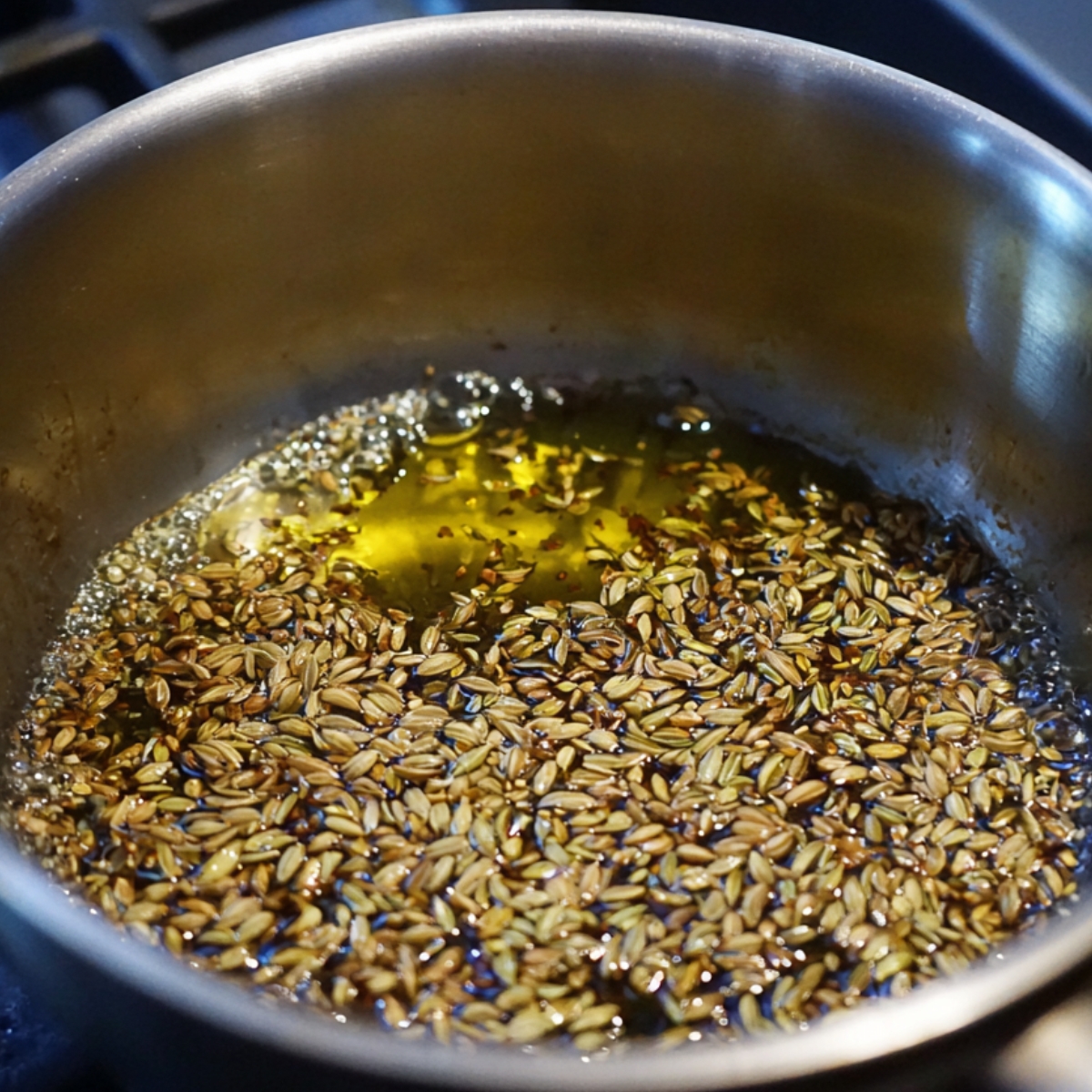 A close-up of fennel seeds frying in hot oil inside a metal pot.