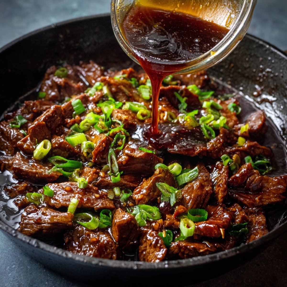“Skillet with tender beef simmering in soy-based sauce, garnished with chopped green onions and being drizzled with more sauce.”