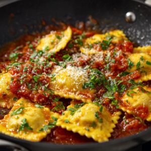 A close-up of ravioli in a skillet, topped with tomato sauce, fresh herbs, and a sprinkle of Parmesan cheese.