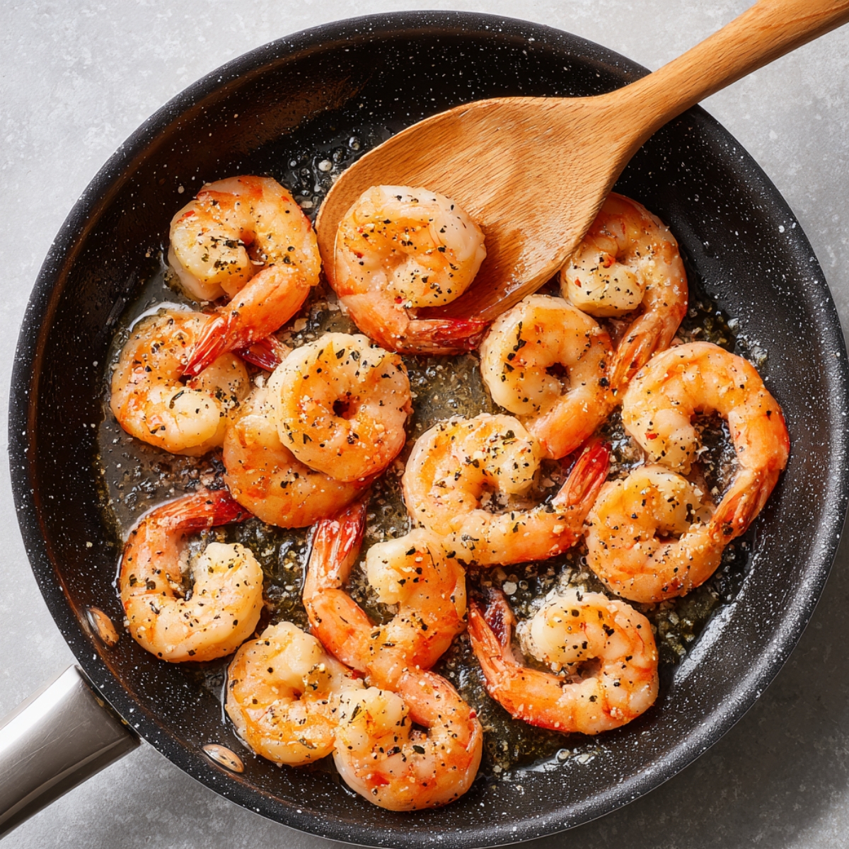 “Shrimp being sautéed in a skillet with melted butter, garlic, and seasonings, stirred with a wooden spoon; shrimp are pink and lightly browned.”