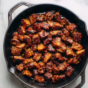 A cast-iron skillet filled with seared beef cubes coated in a glossy brown sauce, garnished with bits of garlic and chili.