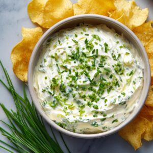 A creamy onion dip garnished with chopped chives, served with crispy potato chips on the side.