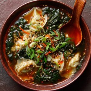 A bowl of spicy potsticker soup topped with wilted greens, sesame seeds, shredded carrots, and green onions, served with a wooden spoon in a rustic brown bowl.