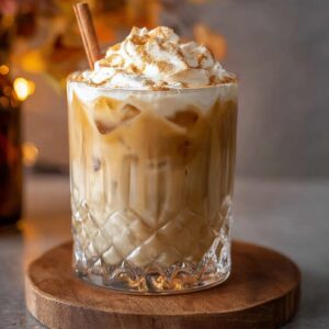 A glass of iced pumpkin spice White Russian topped with whipped cream, sprinkled with cinnamon, and garnished with a cinnamon stick, placed on a wooden coaster.