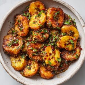 "A bowl of fried plantains with a spicy sauce, garnished with cilantro."