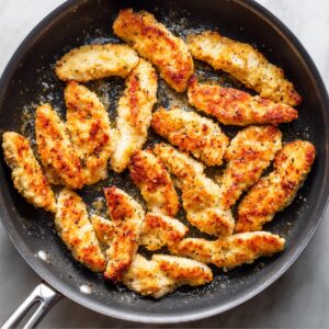 A frying pan filled with crispy golden chicken tenders coated in light breading, cooking in oil.