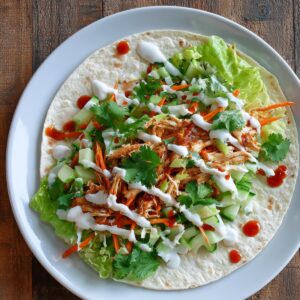 An open tortilla wrap on a white plate with shredded chicken, lettuce, cucumber, carrots, cilantro, and drizzles of white sauce and sweet chili sauce.
