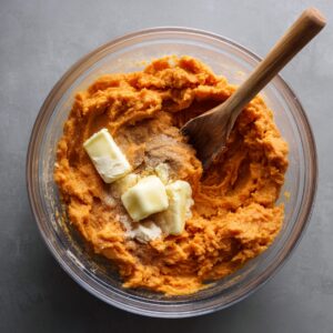 Bowl of mashed sweet potatoes mixed with butter, sugar, and cinnamon, being stirred with a wooden spoon.