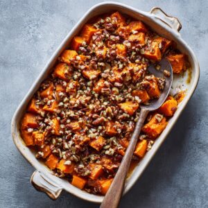 Roasted chunks of sweet potatoes in a casserole dish topped with a brown sugar and pecan crumble, served with a wooden spoon.