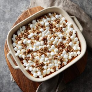 Baked sweet potato casserole topped with toasted mini marshmallows and chopped pecans in a cream-colored baking dish.