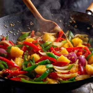 Colorful stir-fry of red and green bell peppers, onions, and pineapple chunks cooking in a wok, releasing steam while being mixed with a wooden spoon.