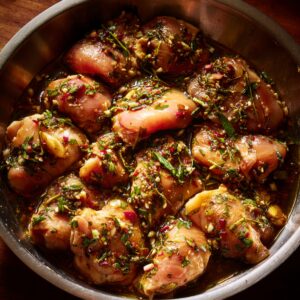Raw chicken pieces marinating in a metal bowl with a mixture of herbs, chopped garlic, chili, and soy-based sauce, prepared for Thai-style fried chicken.