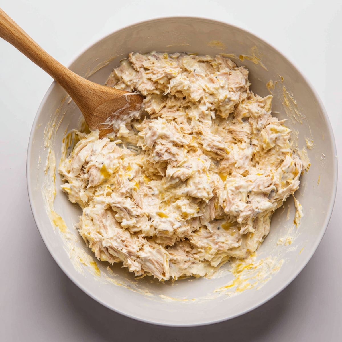 A white bowl containing creamy tuna salad mixed with mayonnaise and a wooden spoon resting inside, showing a thick, smooth consistency.