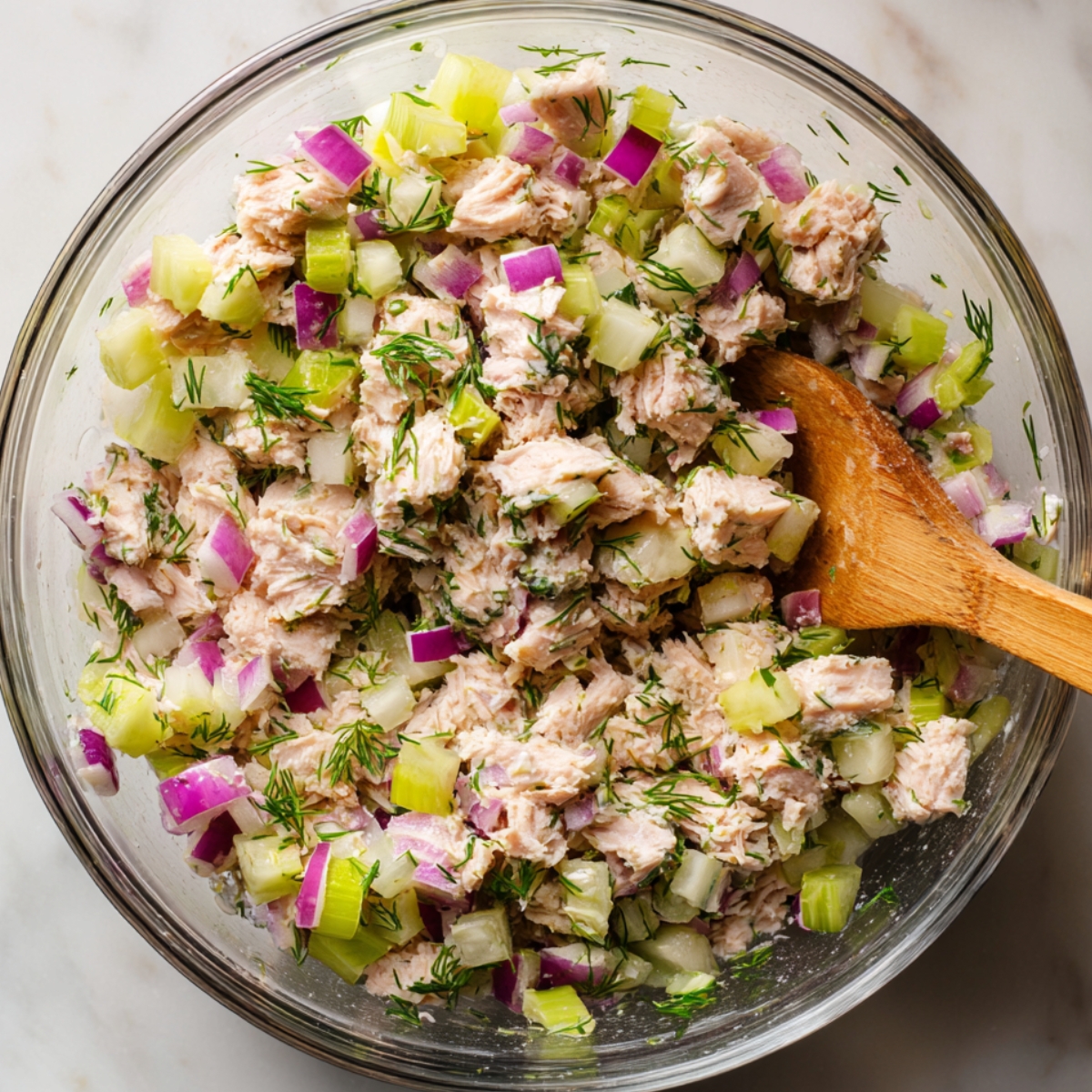 A glass bowl filled with tuna salad containing chunks of tuna, diced celery, red onion, and fresh dill, being mixed with a wooden spoon.