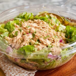 A clear bowl filled with tuna salad made from flaky tuna mixed with chopped lettuce and red onion, lightly dressed, sitting on a wooden table.