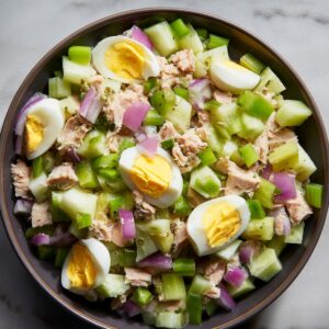A bowl of tuna salad with chopped celery, red onions, and slices of hard-boiled egg on top, placed on a marble surface.