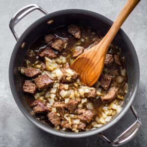 Cubes of beef and diced onions browning in a pot, being stirred with a wooden spoon.