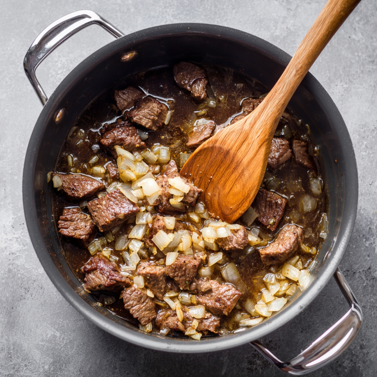 Cubes of beef and diced onions browning in a pot, being stirred with a wooden spoon.