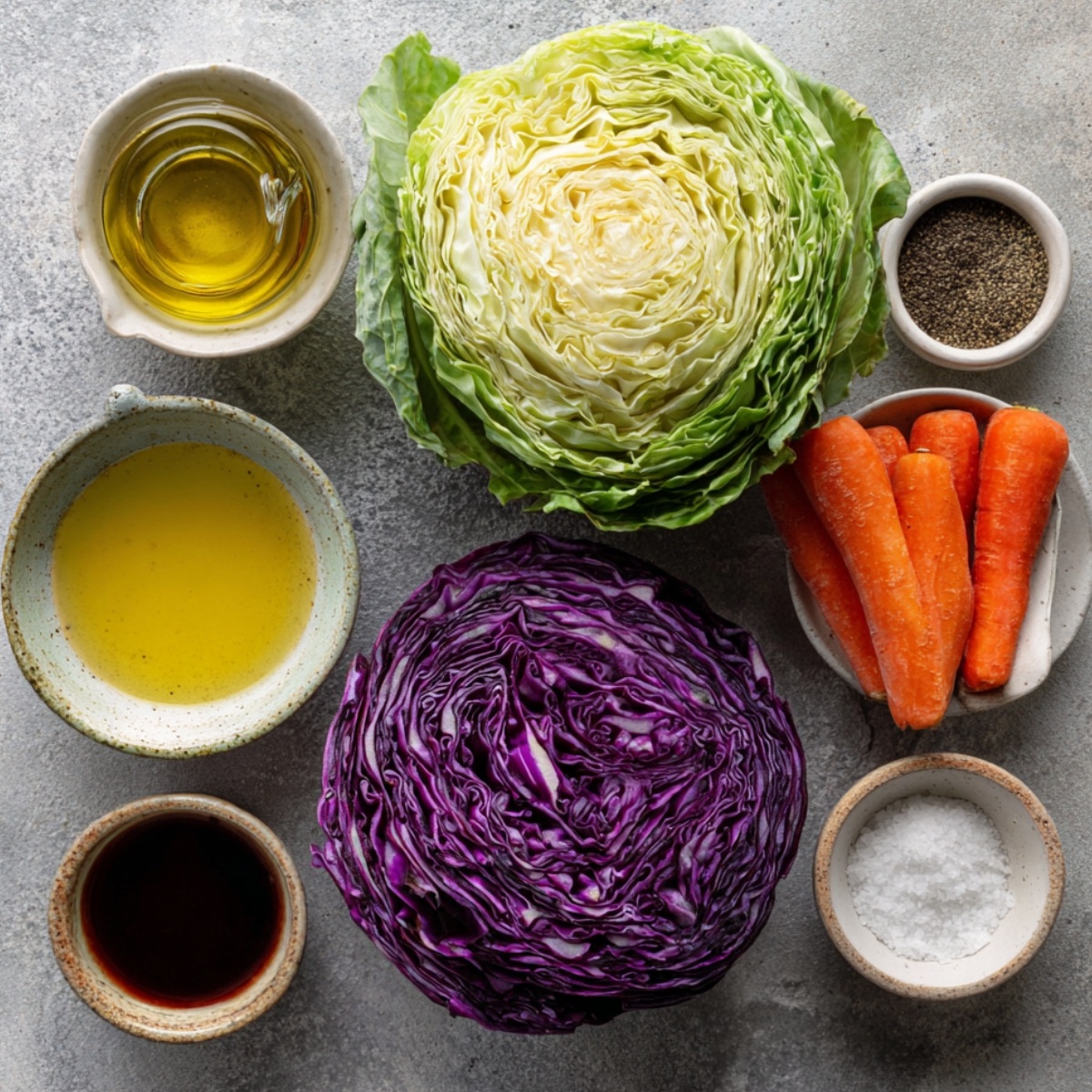 Fresh coleslaw ingredients arranged neatly on a gray surface: green cabbage, purple cabbage, whole carrots, bowls of olive oil, vinegar, salt, and black pepper.