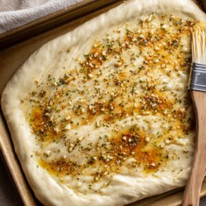 A pan of unbaked pizza dough drizzled with garlic butter and sprinkled with herbs and seasonings.