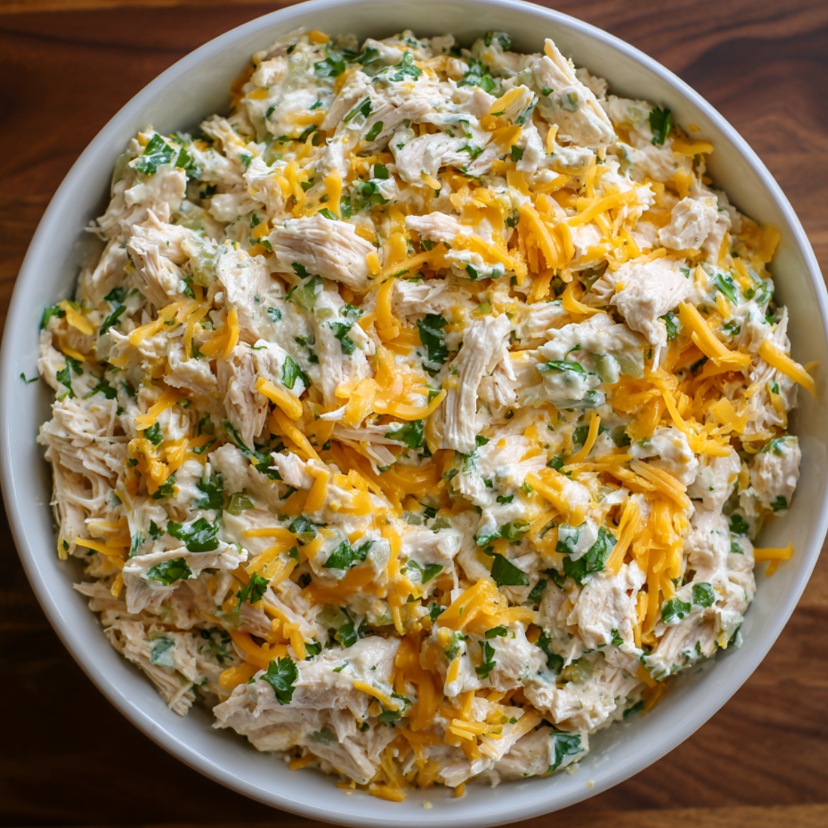 A large bowl filled with shredded chicken mixed with melted cheese, herbs, and creamy sauce, ready to be used as filling for enchiladas.