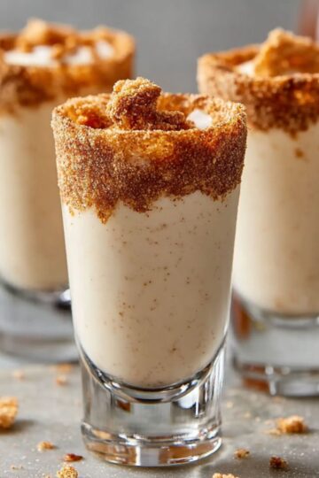 Cinnamon Toast Crunch Shots in clear glass shot glasses, filled with creamy cinnamon liqueur and rimmed with crushed cereal. The drinks sit on a light gray surface sprinkled with crumbs and a few sugar-coated cubes, creating a cozy, real-kitchen vibe.